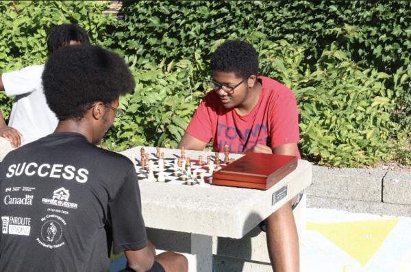 Two black youth are sitting at an outdoor chess table, playing chess.